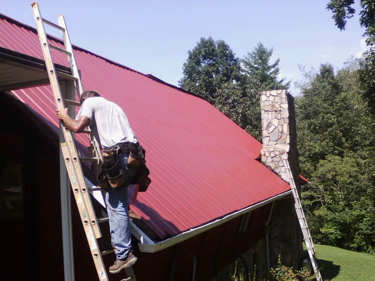 Roofer on a ladder beside a red metal roof and stone chimney — Soffit & Fascia Repair by Roofing Brothers in Donora